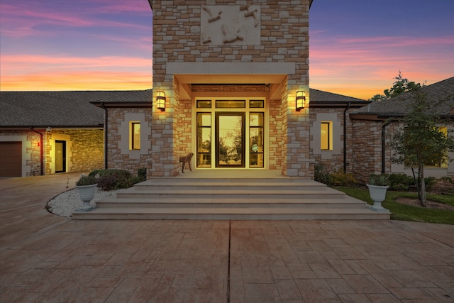 exterior entry at dusk featuring driveway, a shingled roof, an attached garage, and stone siding