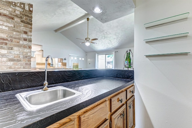 kitchen featuring brown cabinetry, a ceiling fan, dark countertops, lofted ceiling with beams, and a sink