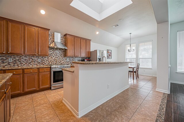 kitchen with brown cabinets, decorative backsplash, appliances with stainless steel finishes, wall chimney range hood, and an island with sink