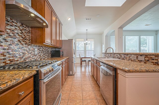 kitchen with light tile patterned floors, appliances with stainless steel finishes, brown cabinetry, a sink, and wall chimney range hood