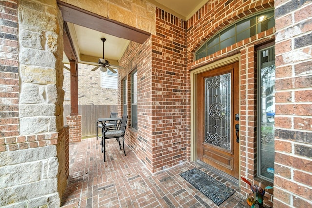 view of exterior entry featuring ceiling fan, brick siding, and fence