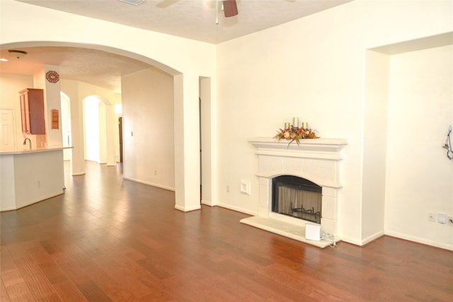 unfurnished living room with a fireplace with raised hearth, ceiling fan, arched walkways, wood finished floors, and baseboards