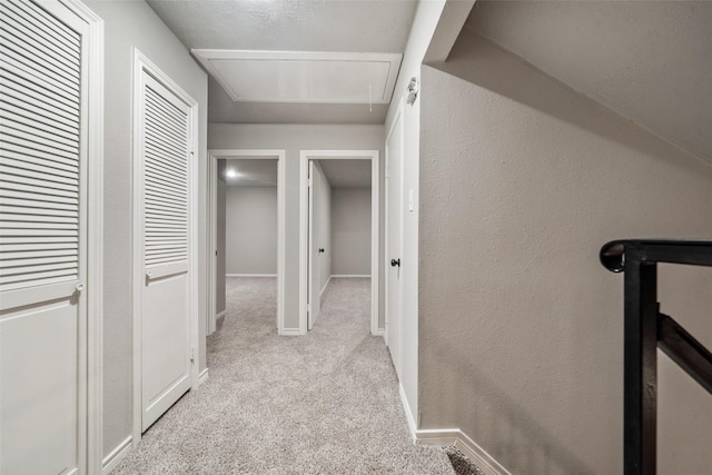 hallway with attic access, light colored carpet, a textured wall, and baseboards