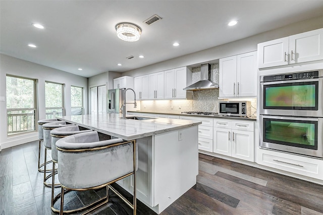 kitchen with a kitchen island with sink, stainless steel appliances, a sink, wall chimney range hood, and tasteful backsplash