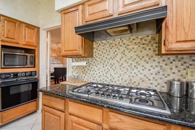 kitchen with dark stone counters, light tile patterned flooring, stainless steel appliances, under cabinet range hood, and tasteful backsplash