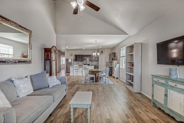 living area featuring ceiling fan with notable chandelier, high vaulted ceiling, light wood-style flooring, and baseboards