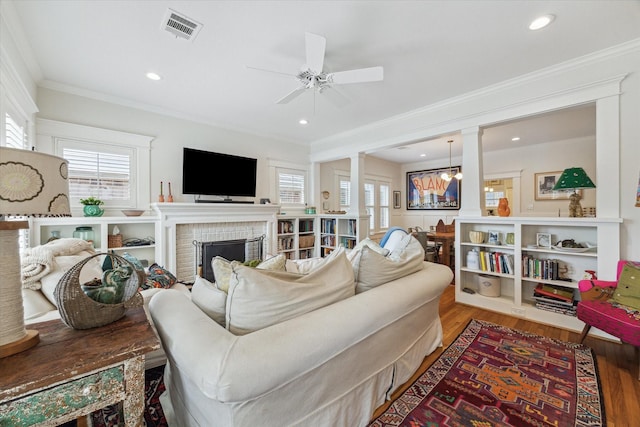 living room with visible vents, wood finished floors, crown molding, a brick fireplace, and ceiling fan with notable chandelier