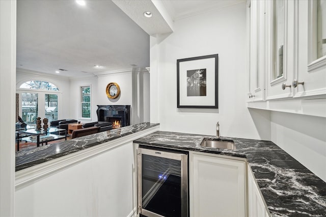 kitchen with a warm lit fireplace, beverage cooler, white cabinetry, ornamental molding, and dark stone counters