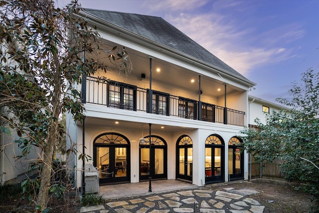 back of house at dusk with a shingled roof, a patio, a balcony, french doors, and stucco siding