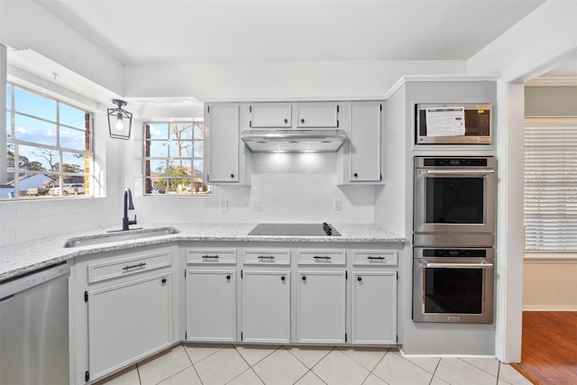 kitchen with under cabinet range hood, stainless steel appliances, a sink, light stone countertops, and tasteful backsplash