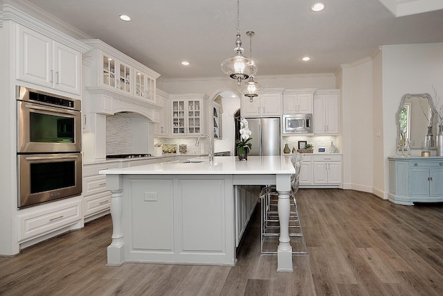 kitchen featuring arched walkways, a sink, light countertops, appliances with stainless steel finishes, and a center island with sink