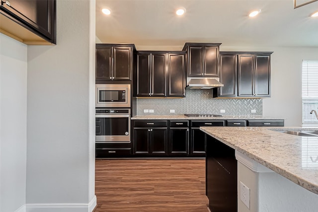 kitchen with light stone counters, under cabinet range hood, a sink, appliances with stainless steel finishes, and backsplash