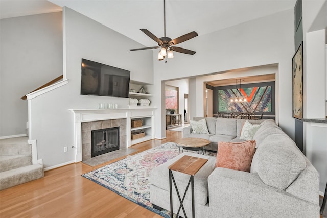 living area with ceiling fan with notable chandelier, wood finished floors, baseboards, stairs, and a tiled fireplace