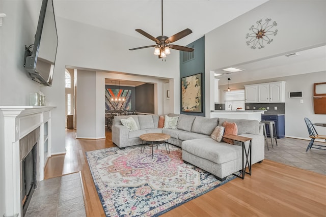 living room with light wood-type flooring, a fireplace with flush hearth, a towering ceiling, and ceiling fan with notable chandelier