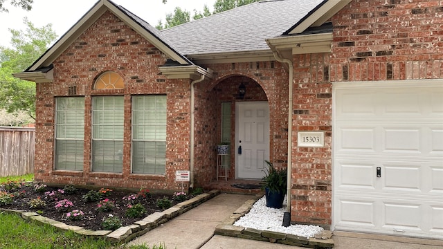 doorway to property featuring brick siding, fence, an attached garage, and roof with shingles