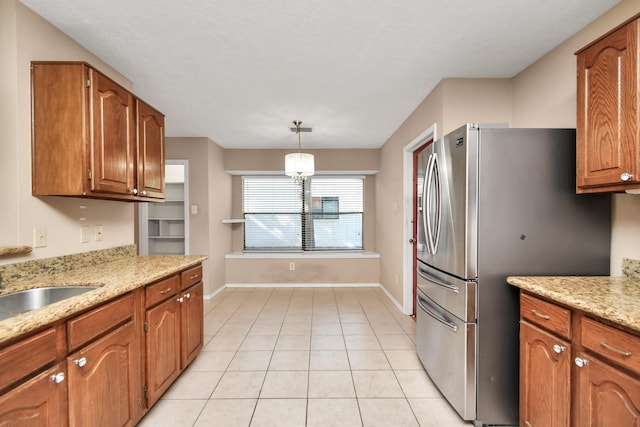 kitchen with light tile patterned floors, brown cabinetry, freestanding refrigerator, hanging light fixtures, and light stone countertops