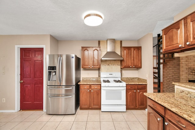 kitchen featuring light stone counters, light tile patterned flooring, white range with gas cooktop, wall chimney range hood, and stainless steel fridge with ice dispenser