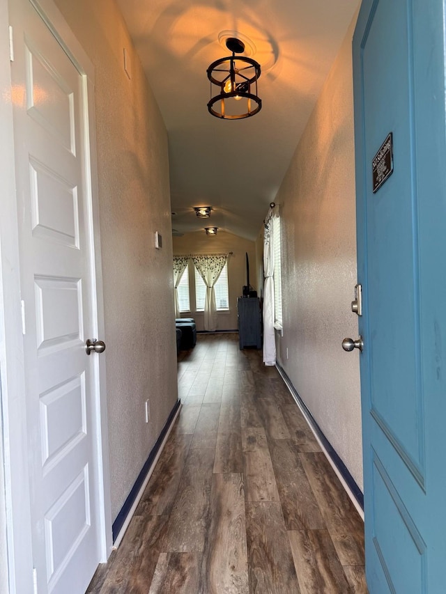 corridor featuring dark wood-style floors, a textured wall, vaulted ceiling, and baseboards