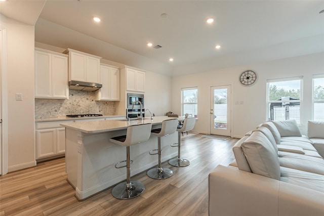 kitchen featuring light wood-type flooring, a center island with sink, backsplash, open floor plan, and light countertops