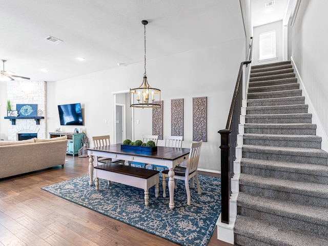 dining area with ceiling fan with notable chandelier, a fireplace, wood finished floors, visible vents, and stairs