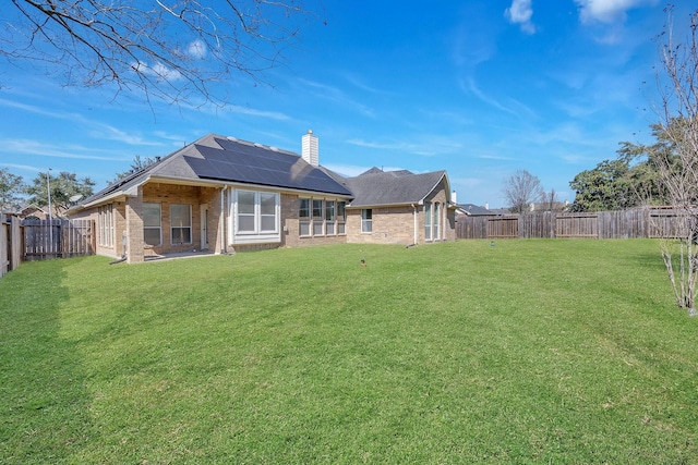 back of house featuring a fenced backyard, brick siding, a yard, roof mounted solar panels, and a chimney