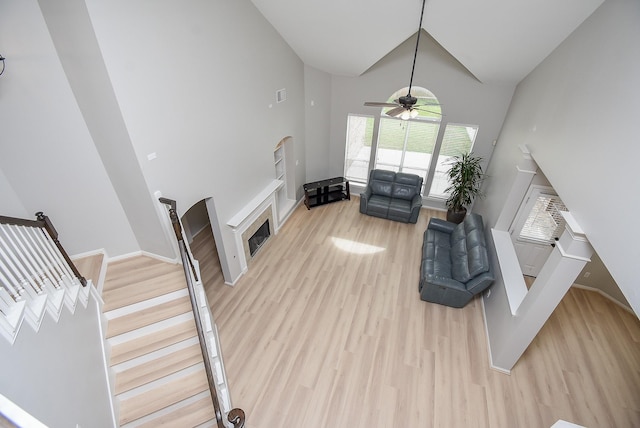 living area featuring high vaulted ceiling, a fireplace, a ceiling fan, stairs, and light wood finished floors