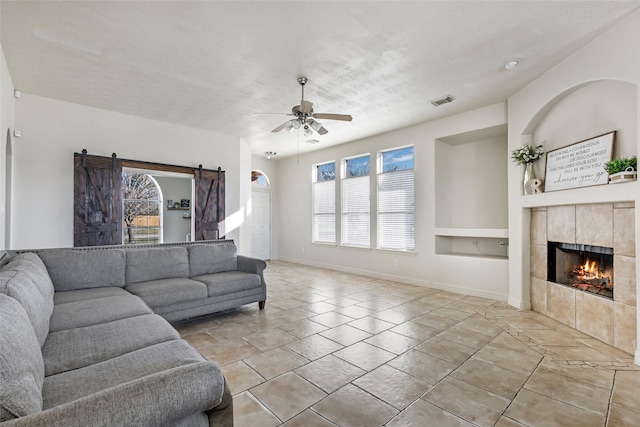 living area featuring a barn door, visible vents, a ceiling fan, a tile fireplace, and built in shelves