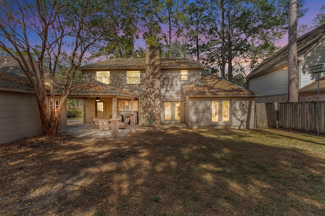 back of house at dusk with french doors, a patio, a chimney, fence, and a yard