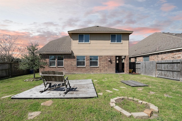 back of house at dusk featuring brick siding, a yard, roof with shingles, a fenced backyard, and a wooden deck