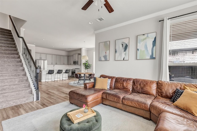 living room with crown molding, light wood finished floors, visible vents, stairway, and ceiling fan