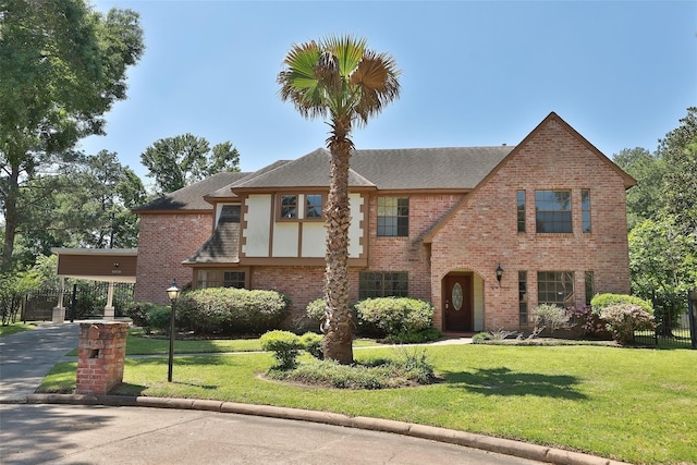 tudor house featuring brick siding, a shingled roof, a front yard, fence, and driveway