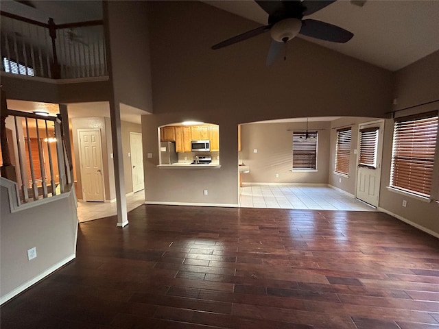 unfurnished living room with high vaulted ceiling, ceiling fan with notable chandelier, baseboards, and wood finished floors