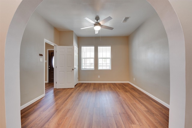 empty room with arched walkways, light wood finished floors, visible vents, ceiling fan, and baseboards