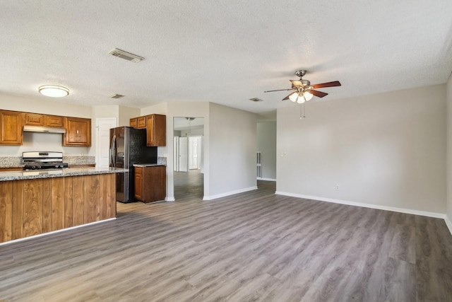 kitchen with under cabinet range hood, stainless steel gas range oven, brown cabinetry, and freestanding refrigerator