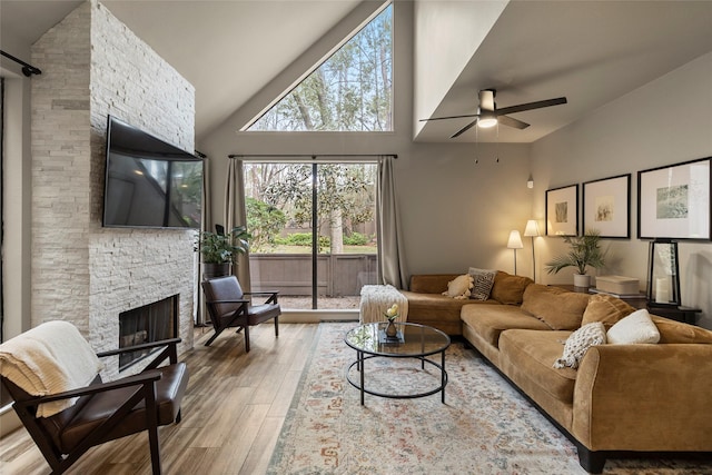 living area with ceiling fan, high vaulted ceiling, a stone fireplace, and wood finished floors