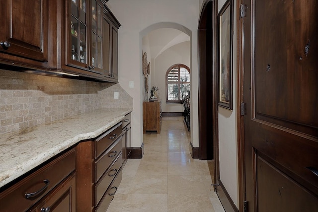 kitchen featuring arched walkways, light stone counters, dark brown cabinets, backsplash, and glass insert cabinets