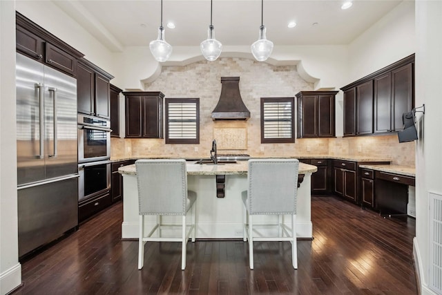 kitchen featuring tasteful backsplash, an island with sink, wall chimney exhaust hood, appliances with stainless steel finishes, and dark brown cabinets