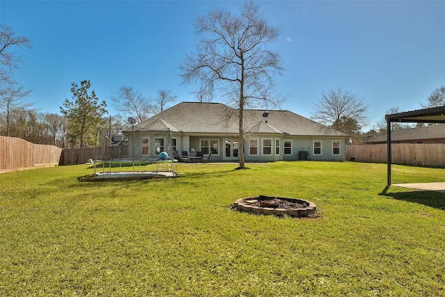 rear view of house with a trampoline, a fenced backyard, a lawn, and a fire pit