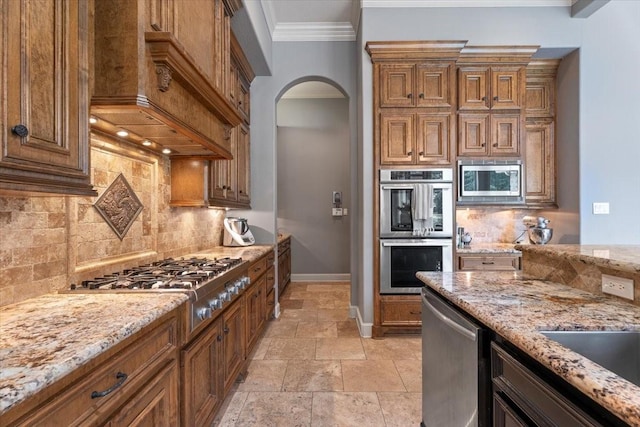 kitchen featuring stone tile floors, brown cabinetry, arched walkways, ornamental molding, and stainless steel appliances