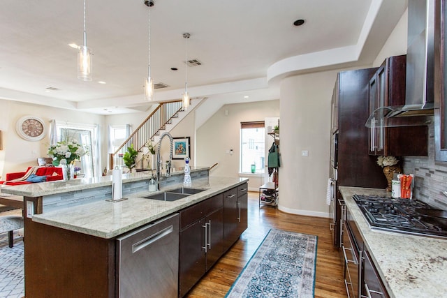 kitchen featuring an island with sink, a sink, dark brown cabinetry, appliances with stainless steel finishes, and wall chimney exhaust hood