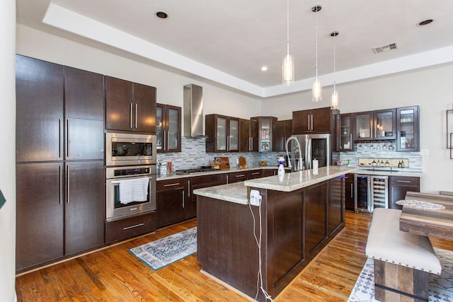 kitchen with beverage cooler, a tray ceiling, wood finished floors, stainless steel appliances, and wall chimney range hood