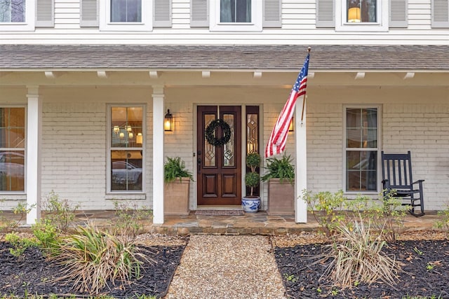 entrance to property featuring a shingled roof, a porch, and brick siding