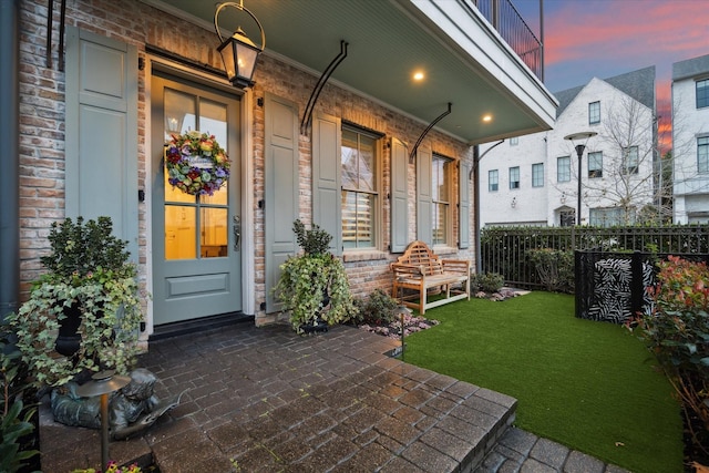 exterior entry at dusk with brick siding, a lawn, a porch, and fence