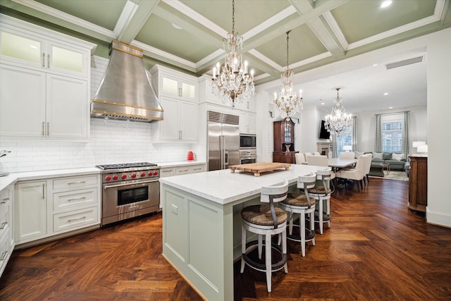 kitchen featuring built in appliances, a kitchen island, white cabinetry, wall chimney range hood, and an inviting chandelier