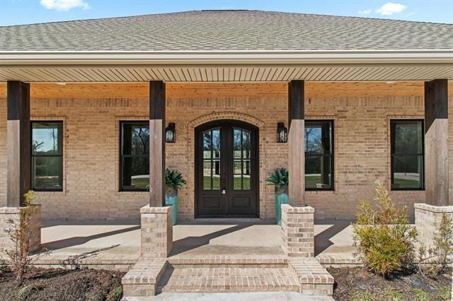 view of exterior entry with a shingled roof, a porch, and brick siding