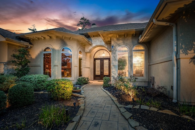 exterior entry at dusk with a shingled roof, french doors, and stucco siding