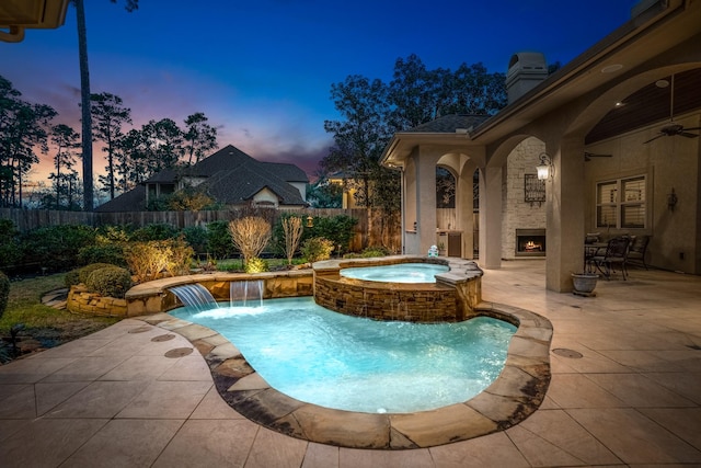 pool at dusk featuring a warm lit fireplace, an in ground hot tub, fence, a ceiling fan, and a patio area