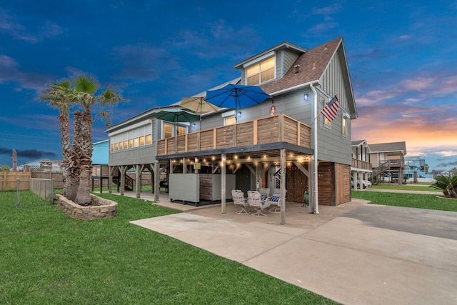 back of house at dusk featuring a yard, a patio, a shingled roof, board and batten siding, and a deck