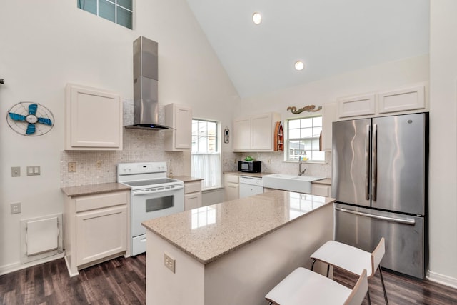 kitchen with white appliances, a center island, wall chimney range hood, white cabinetry, and a sink