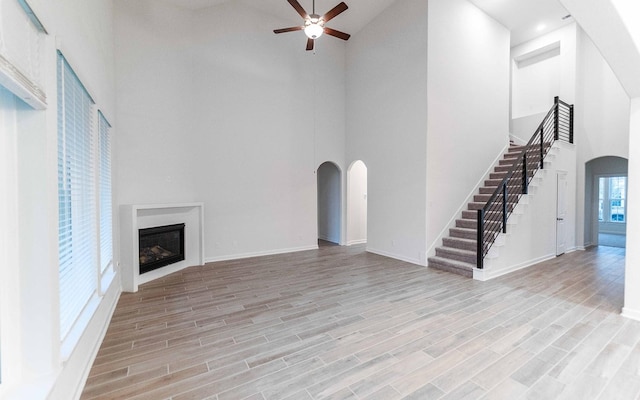 unfurnished living room with arched walkways, light wood finished floors, a glass covered fireplace, ceiling fan, and stairs